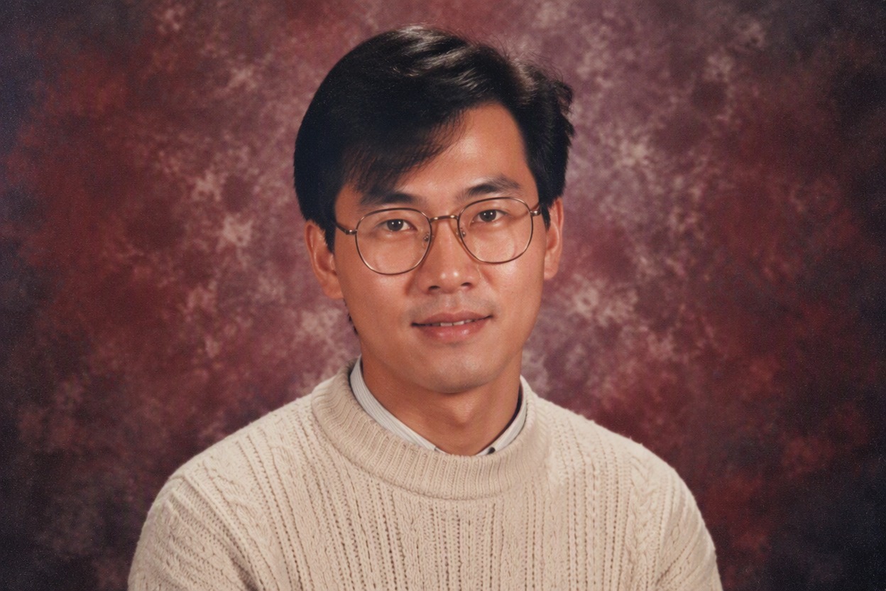 One fictional adult Black man with short sculpted high-top hair, teal patterned knit sweater and charcoal blazer posed for an early-1990s school yearbook portrait against a slate-toned studio backdrop